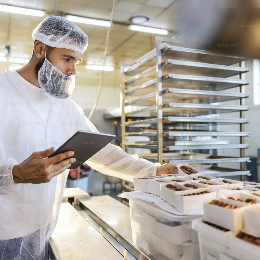 Male bakery worker in a hairnet and protective suit inspecting chocolates on a tray while holding a tablet in a pastry kitchen.